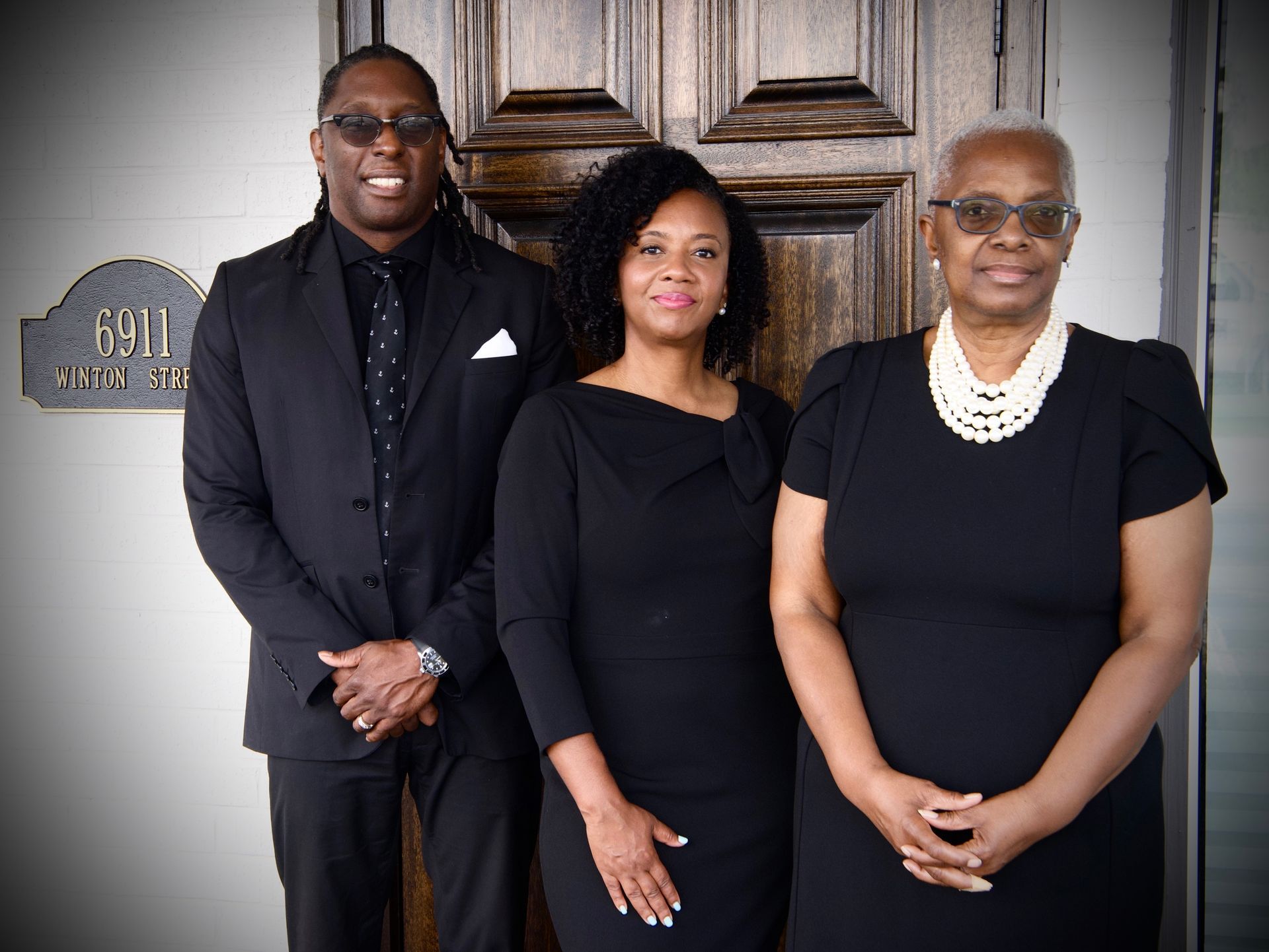 Three people in black outfits pose in front of a wooden door, smiling at the camera.