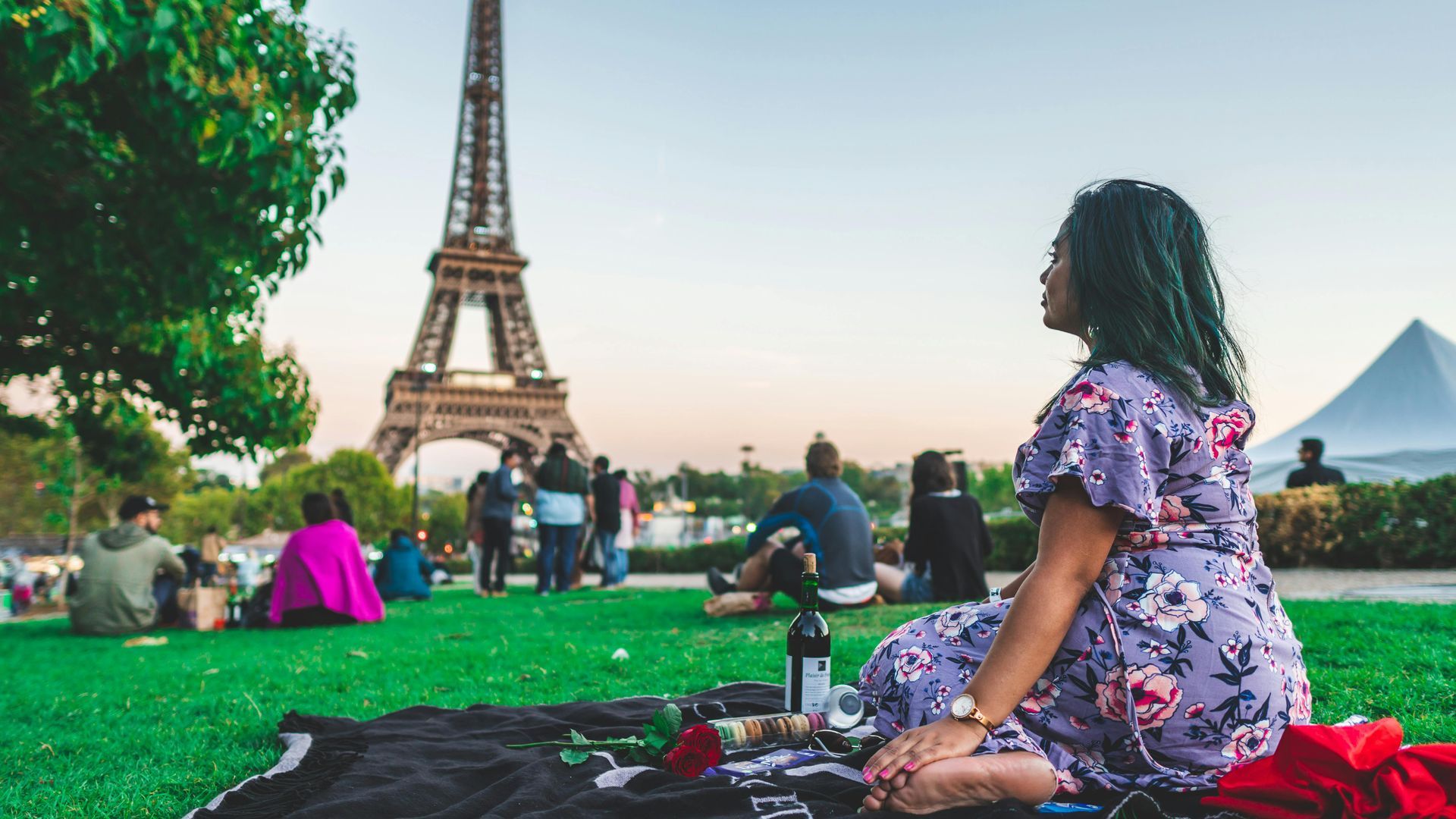 Woman seated on a blanket in front of the Eiffel Tower, enjoying a picnic on a grassy lawn.