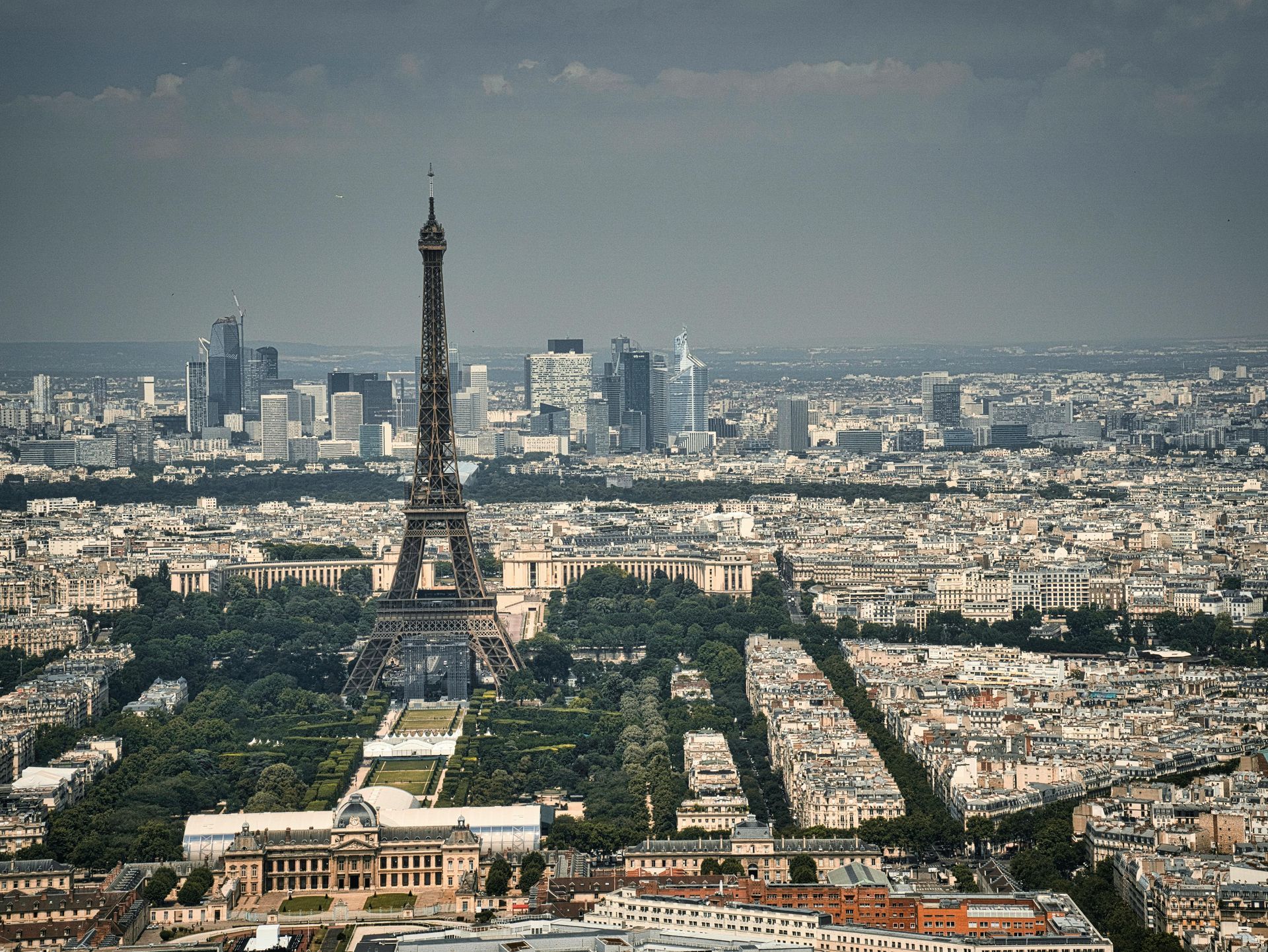 Eiffel Tower over Paris skyline. Grey sky. Buildings, trees, and city streets in view.