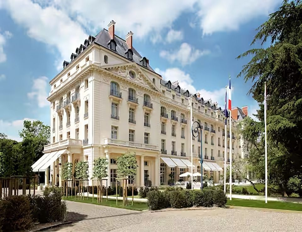 Elegant light-colored hotel building with multiple stories and balconies, set in a park-like area with trees and flags.