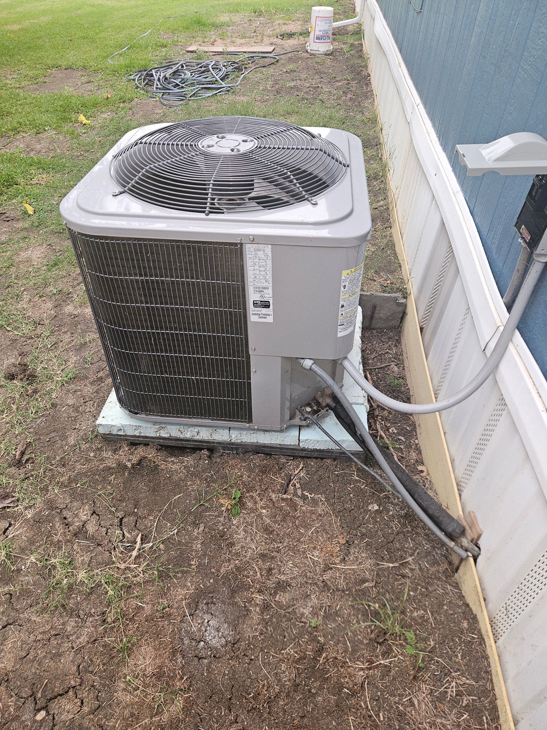 Outdoor air conditioner unit beside a house, with grass and gravel around it and cables attached.