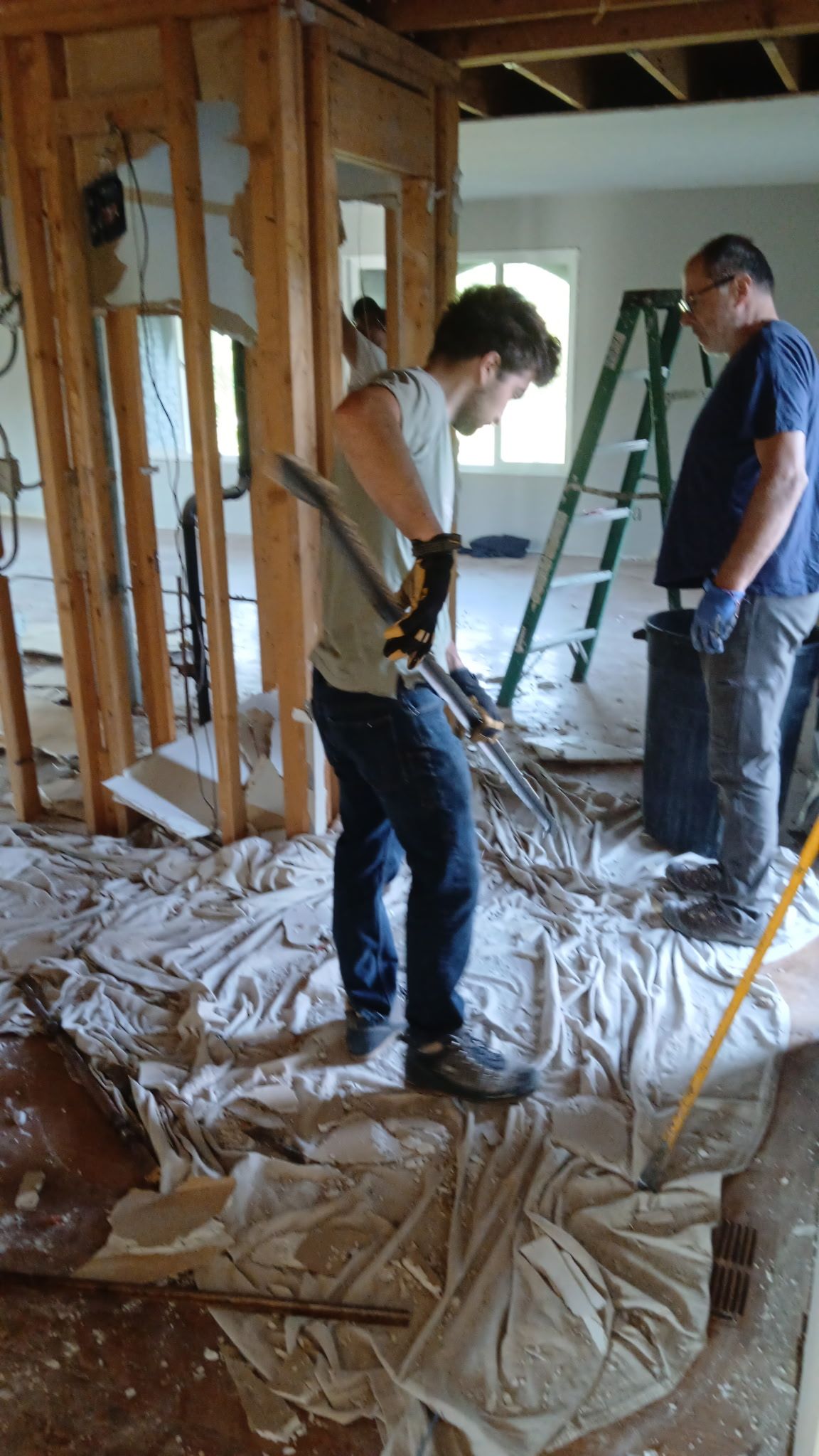 Two workers standing in a room under renovation with exposed studs and debris-covered floor