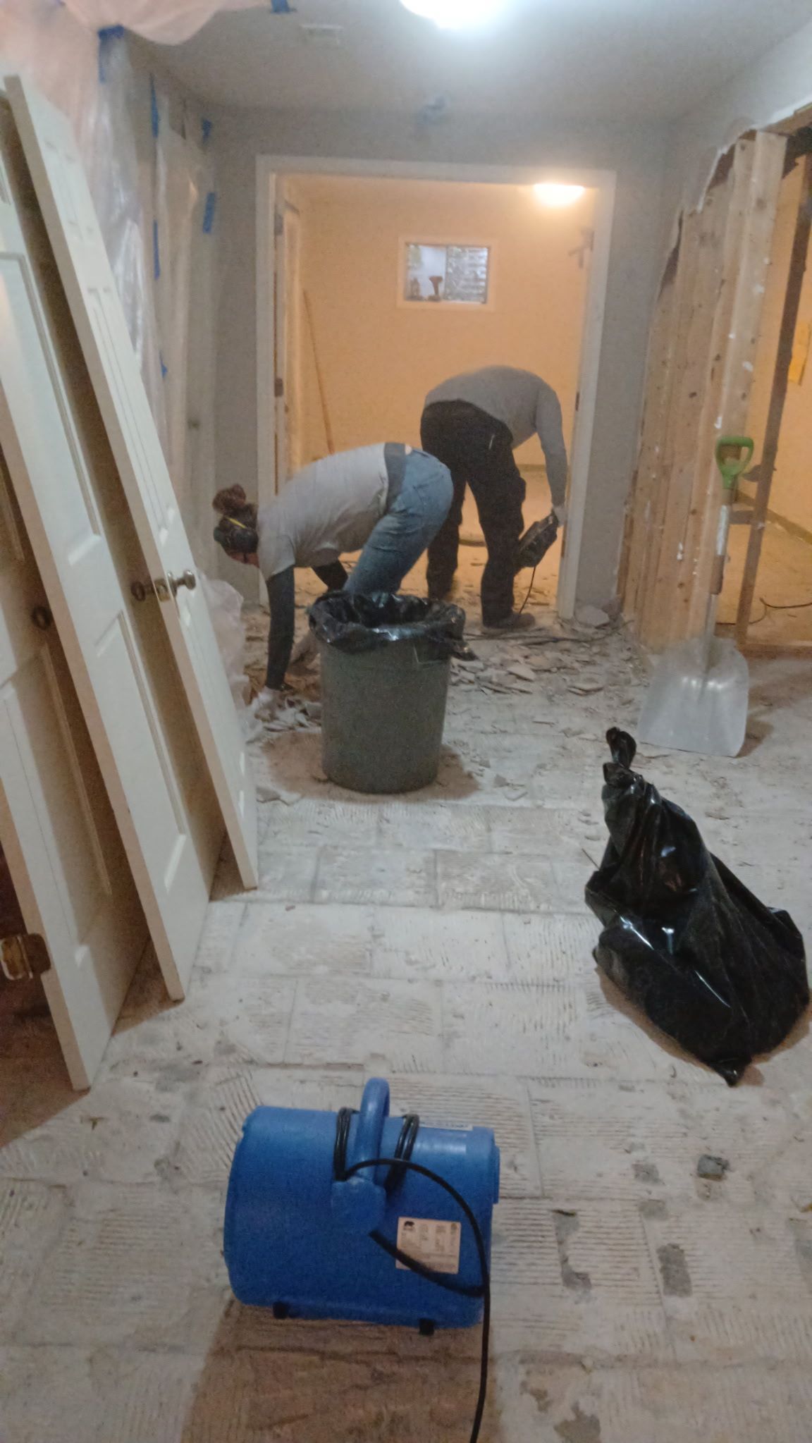 Two workers bend over in a dusty hallway with tools, boards, and renovation debris scattered around.