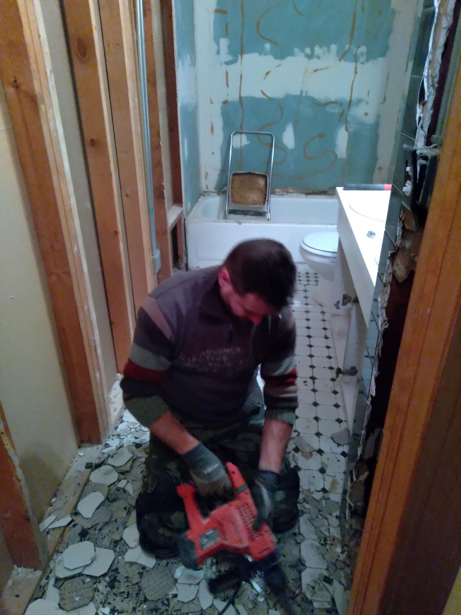 Worker using a red power tool to remove floor tiles in a narrow bathroom under renovation