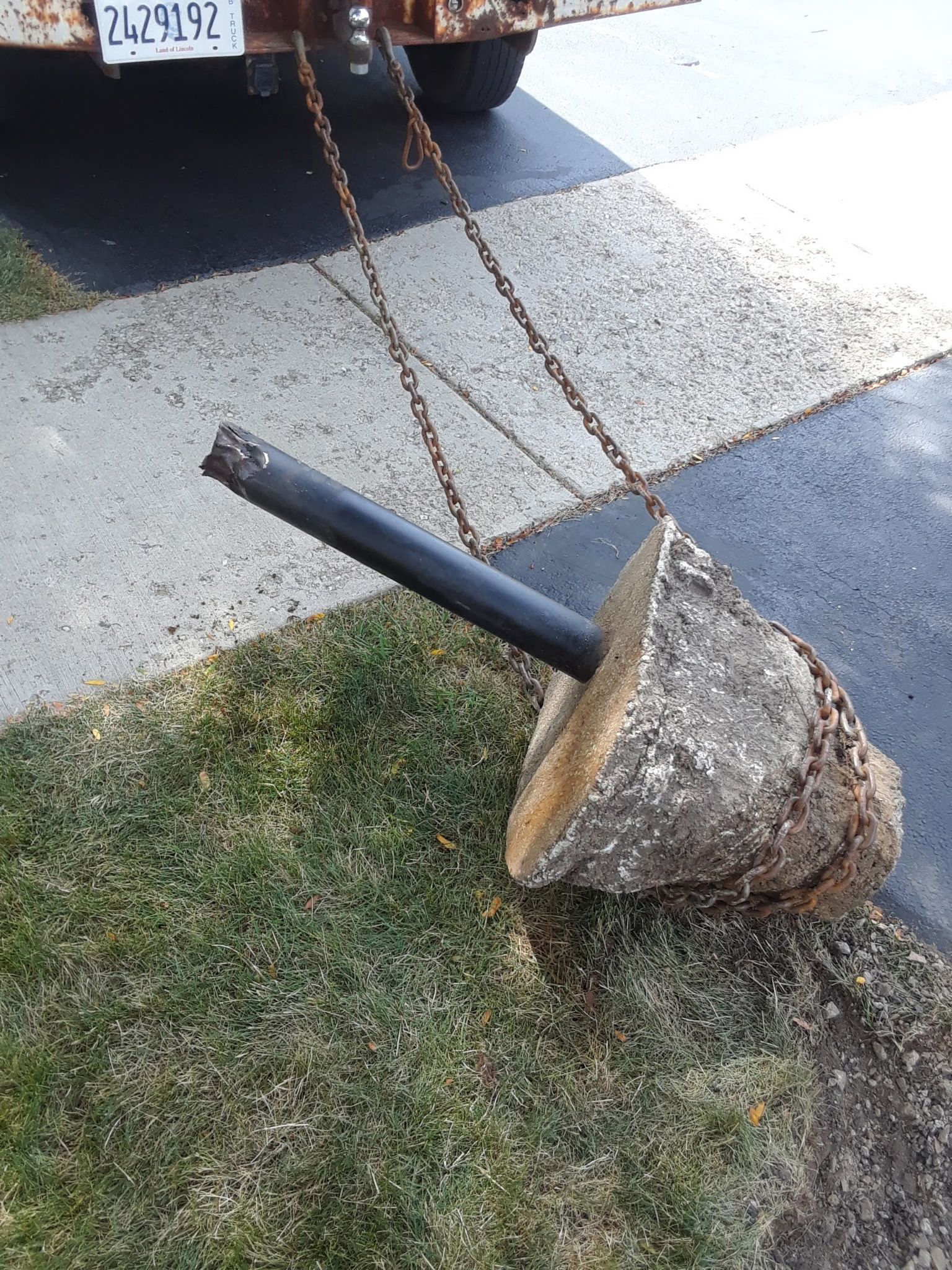 Concrete block with a black metal pipe and chains attached, resting on grass beside a driveway.