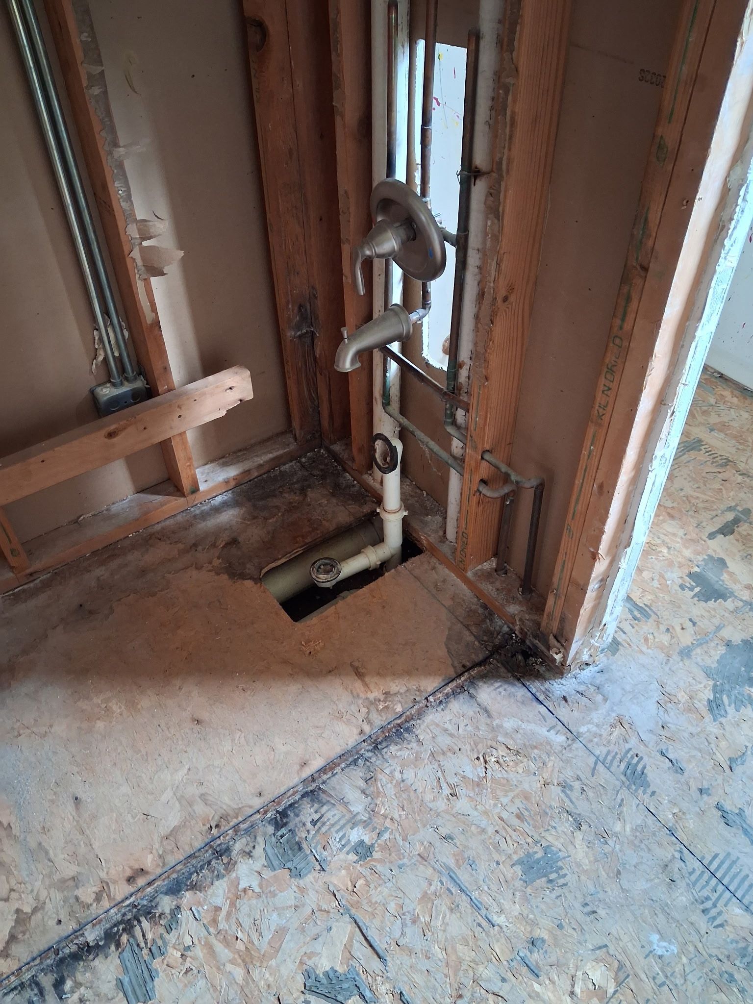 Exposed plumbing pipes and a drain opening inside an unfinished wall with dusty flooring.