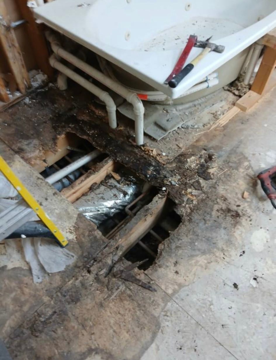 Bathroom floor with a large broken hole exposing pipes near a white tub and tools nearby