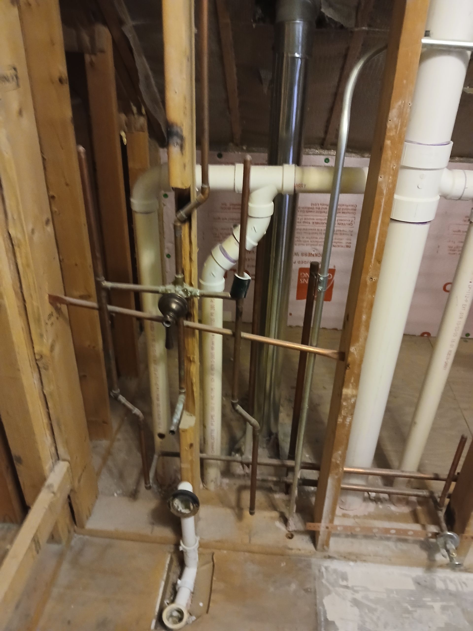 Exposed plumbing and pipes inside an unfinished wall with wooden framing and insulation