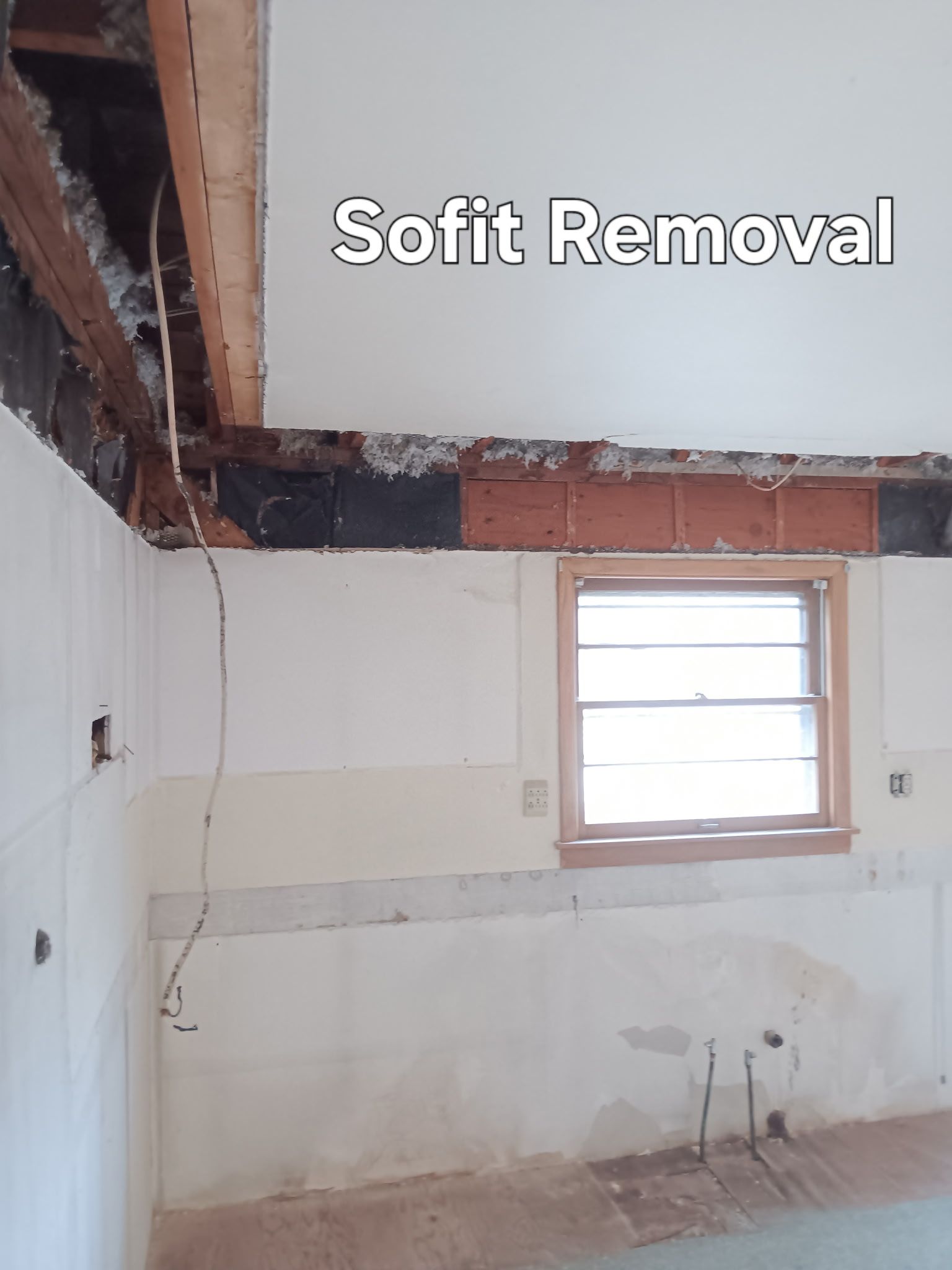 Partially demolished room with exposed framing, white wall panels, and a window; text reads “Soffit Removal”