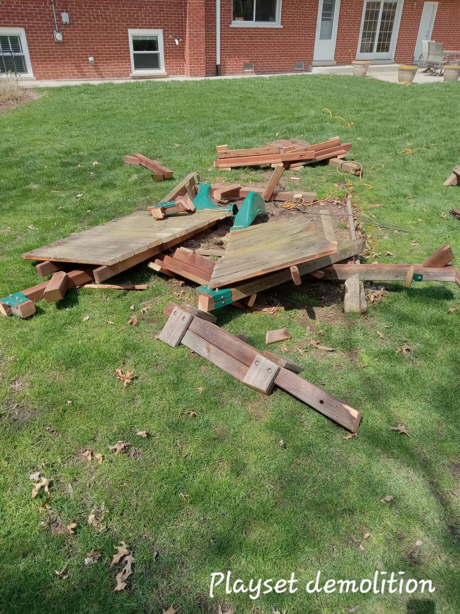 Broken wooden playground structure in a grassy yard near a brick building, with debris scattered around.