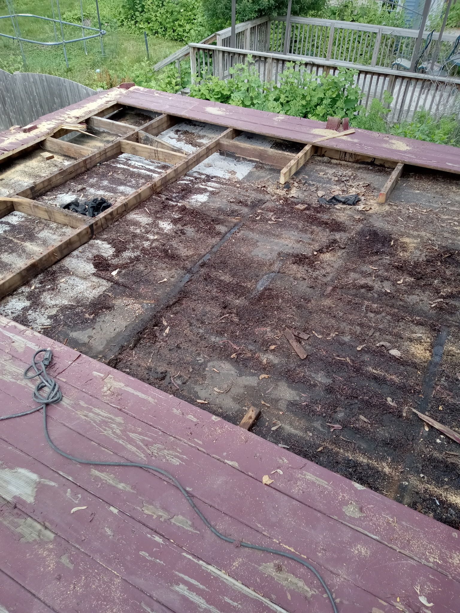 Rotting wooden deck boards removed, exposing soil and joists beside a fenced yard.