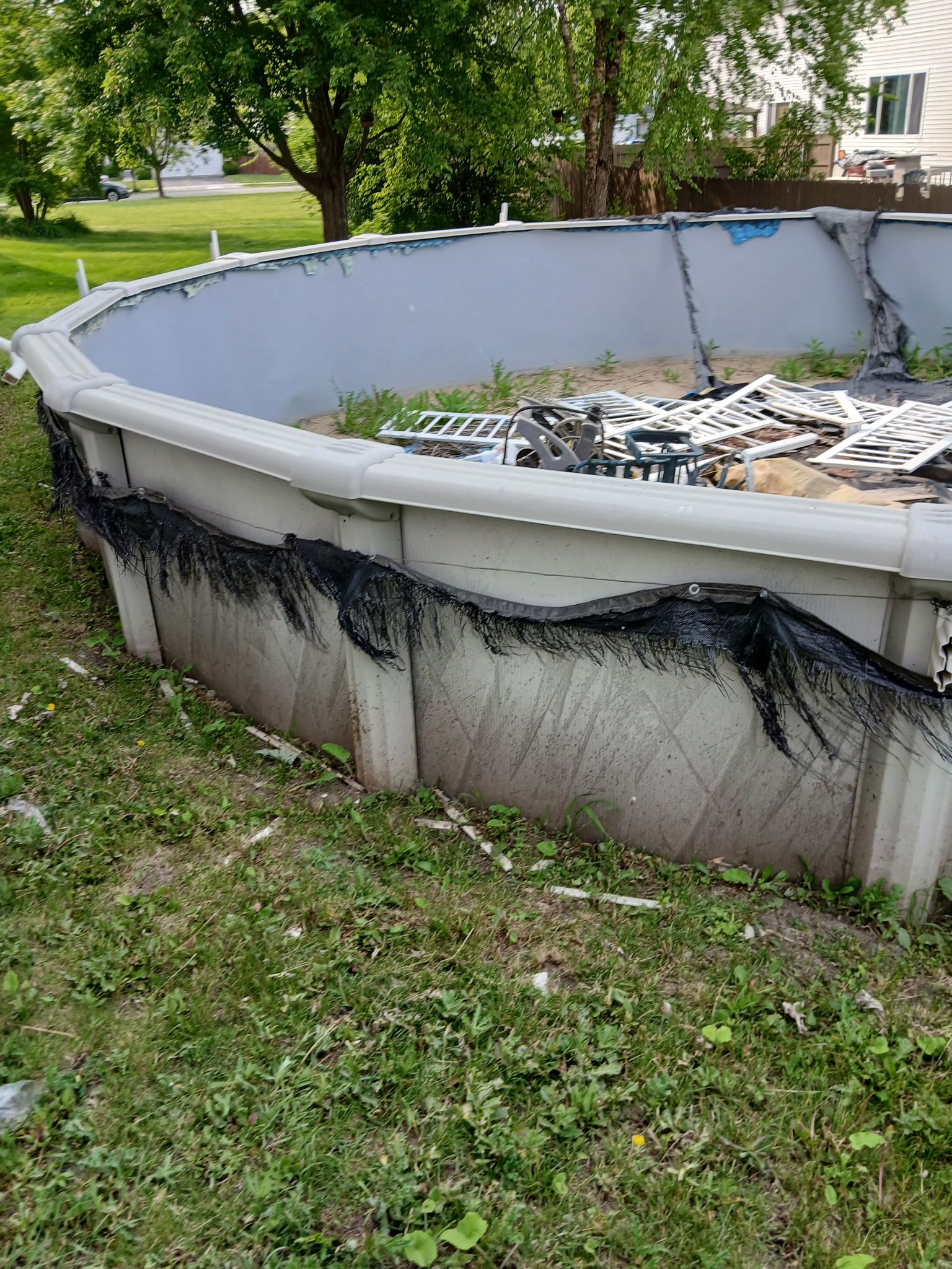 Above-ground pool with damaged black liner and debris in a grassy backyard