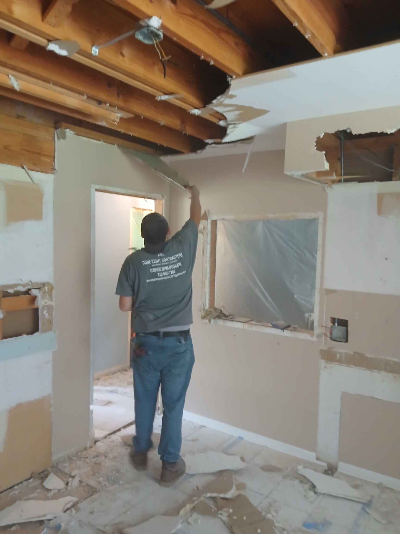 Worker installing drywall in a partially demolished room with exposed ceiling beams and debris on the floor