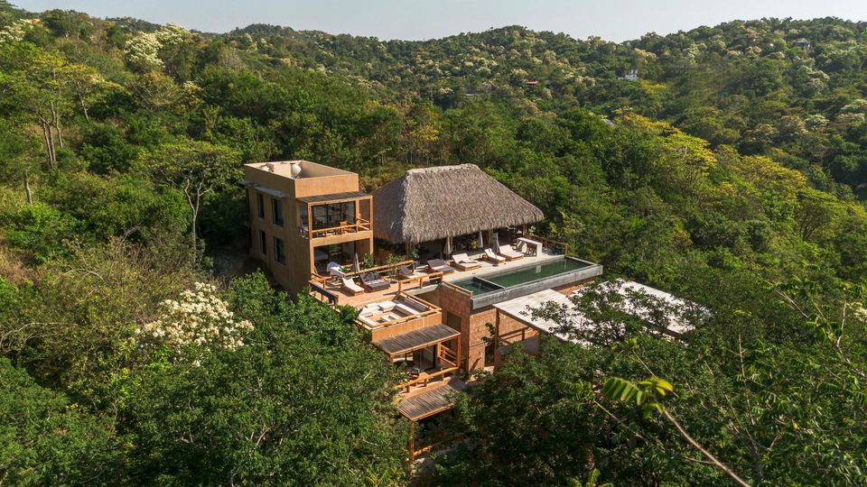 A modern lodge with a thatched roof nestled in dense green forested hills.