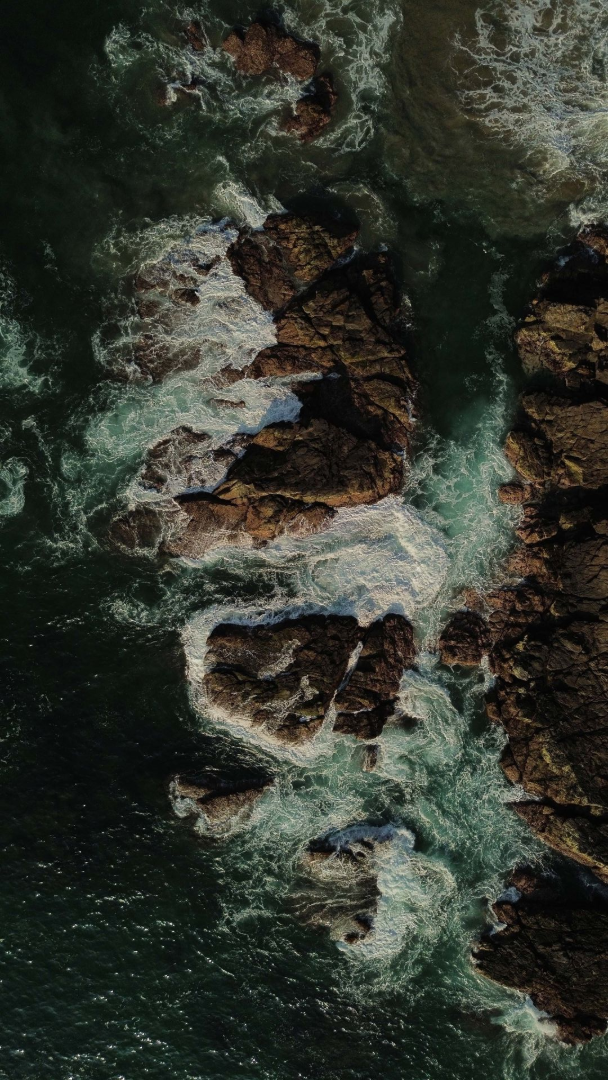 Aerial view of waves crashing against dark rocky shoreline and turquoise water