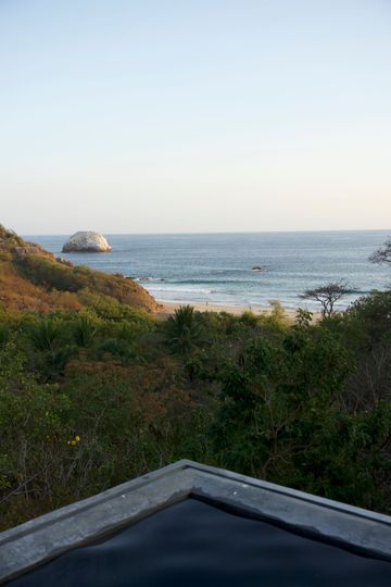Coastal view at sunset with ocean, rocky shore, and green shrubs in the foreground