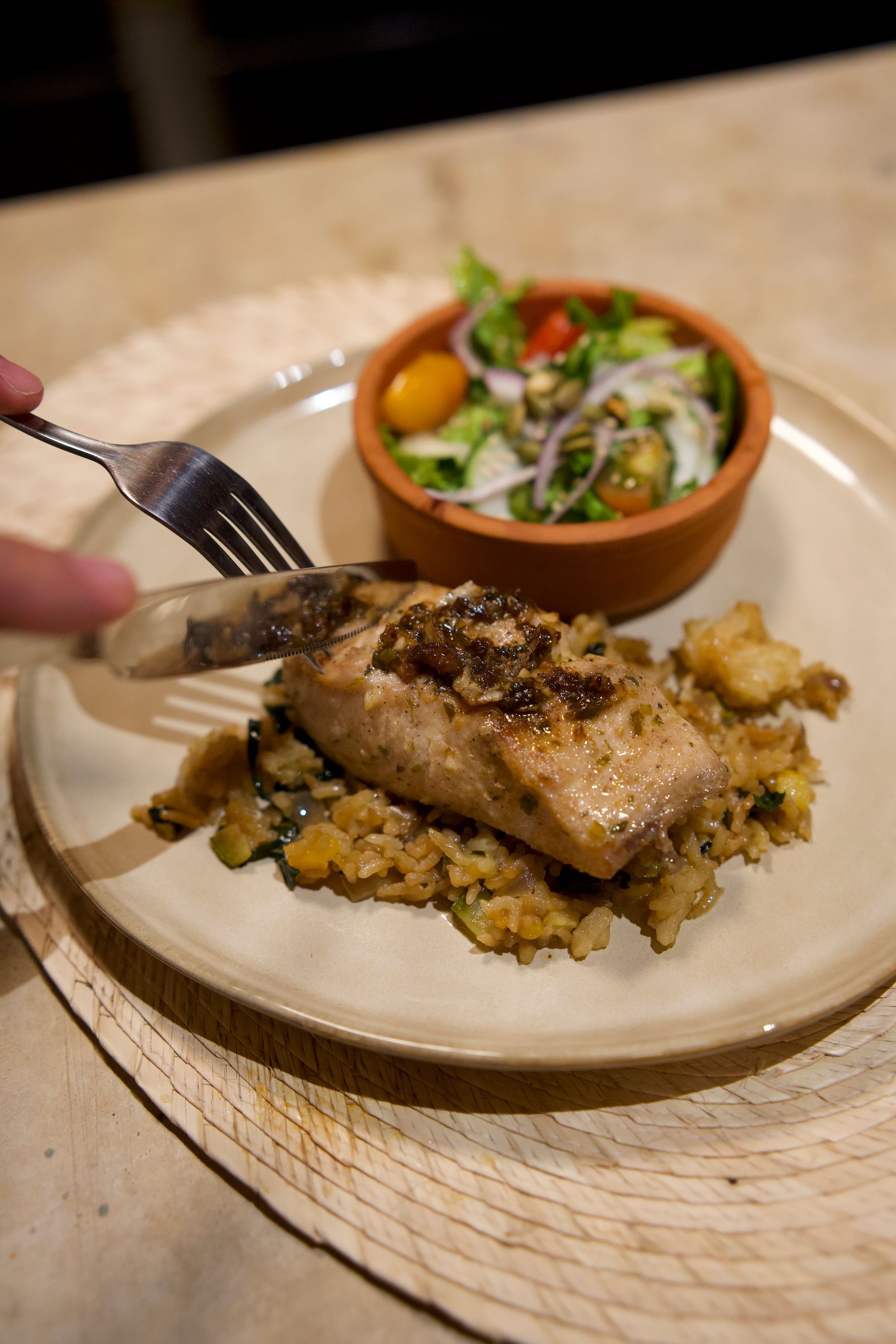 Plated grilled fish over rice with a small salad bowl on a table, with a fork nearby.