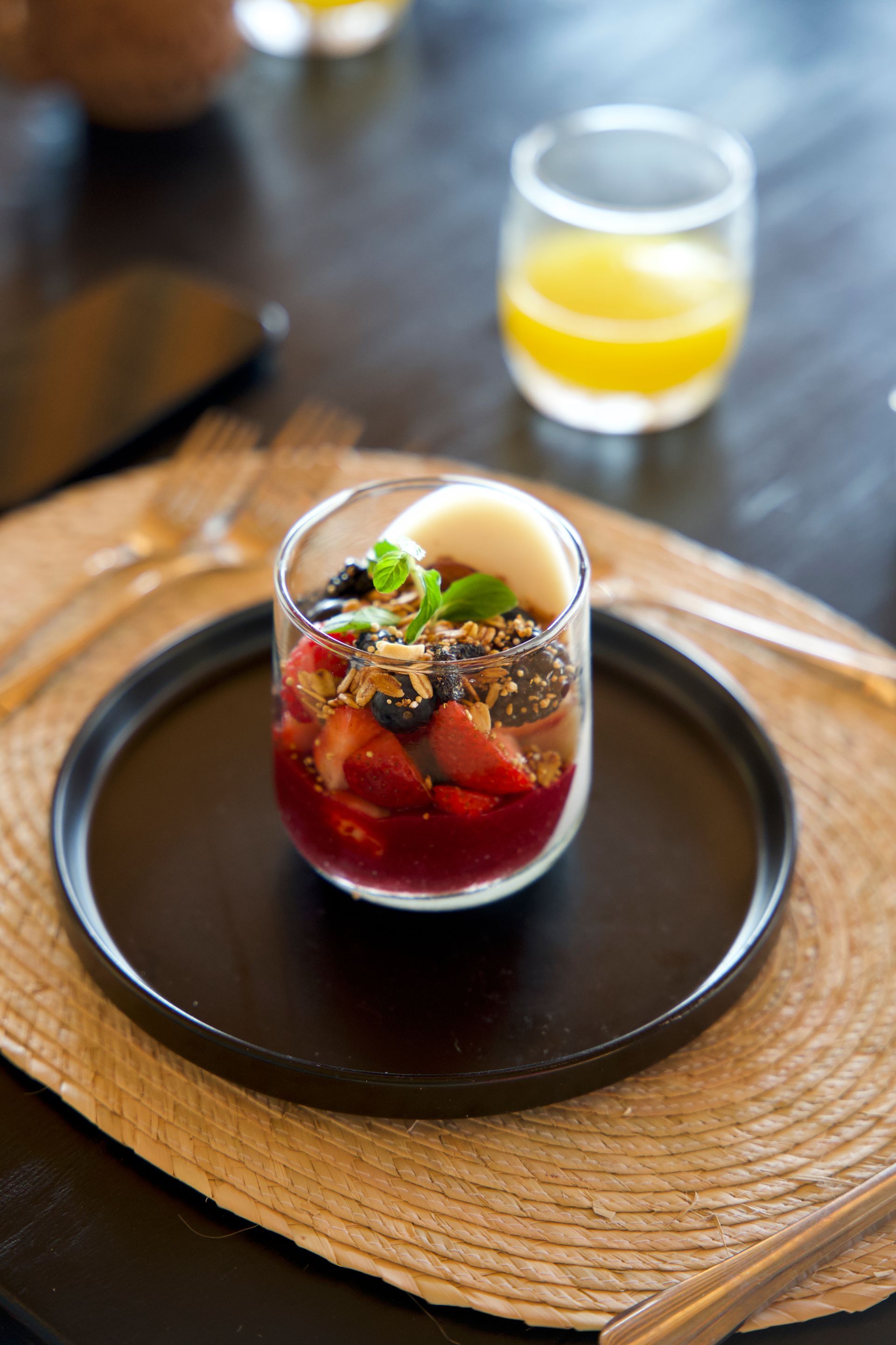 Dessert parfait in a glass on a black plate, with a woven placemat and a yellow drink in the background
