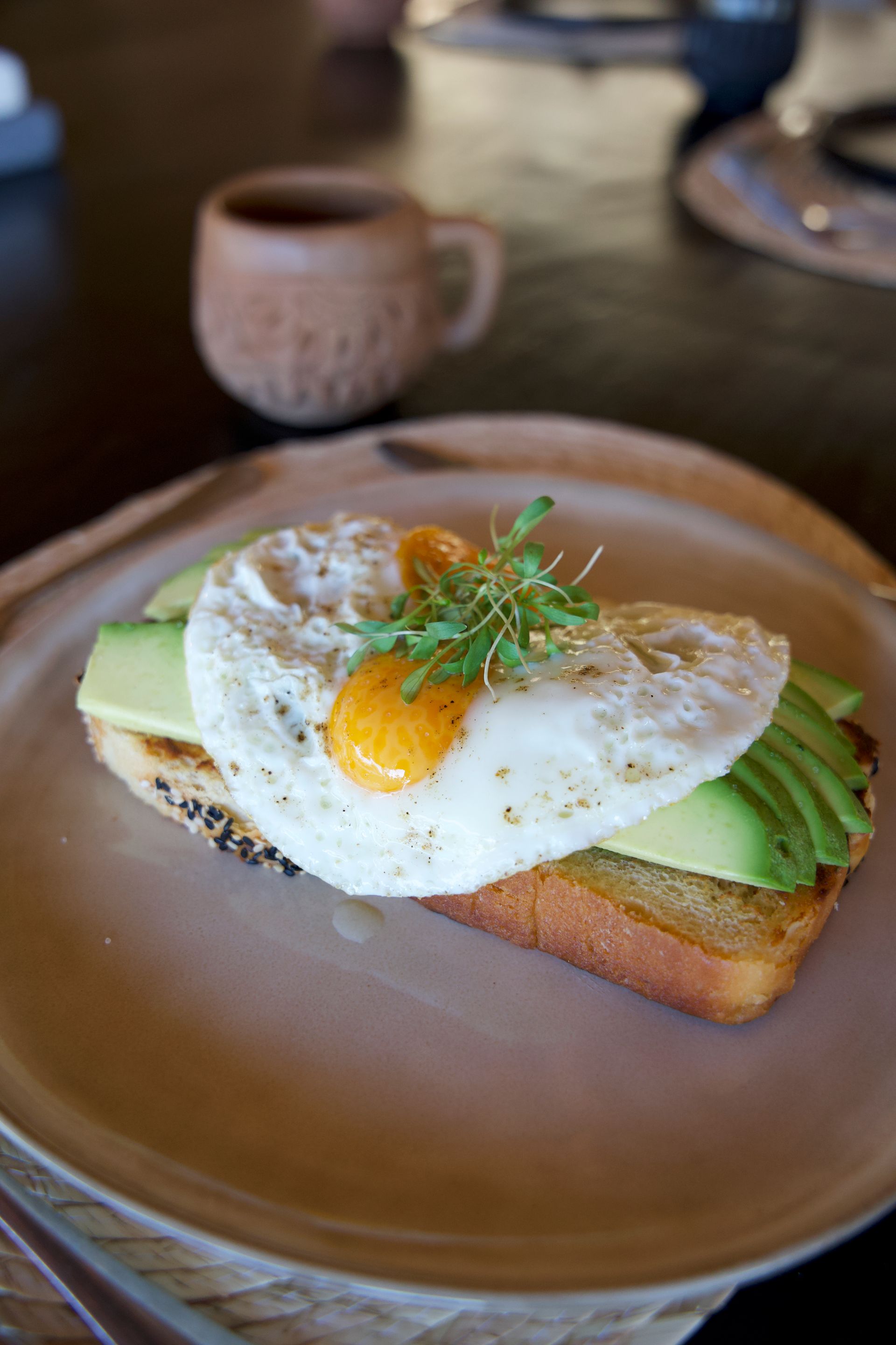 Fried egg and avocado toast on a tan plate, with a coffee cup in the background