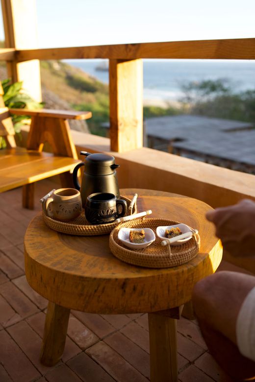 Tea set on a wooden table on a sunlit balcony, with pastries and a person’s hand nearby.