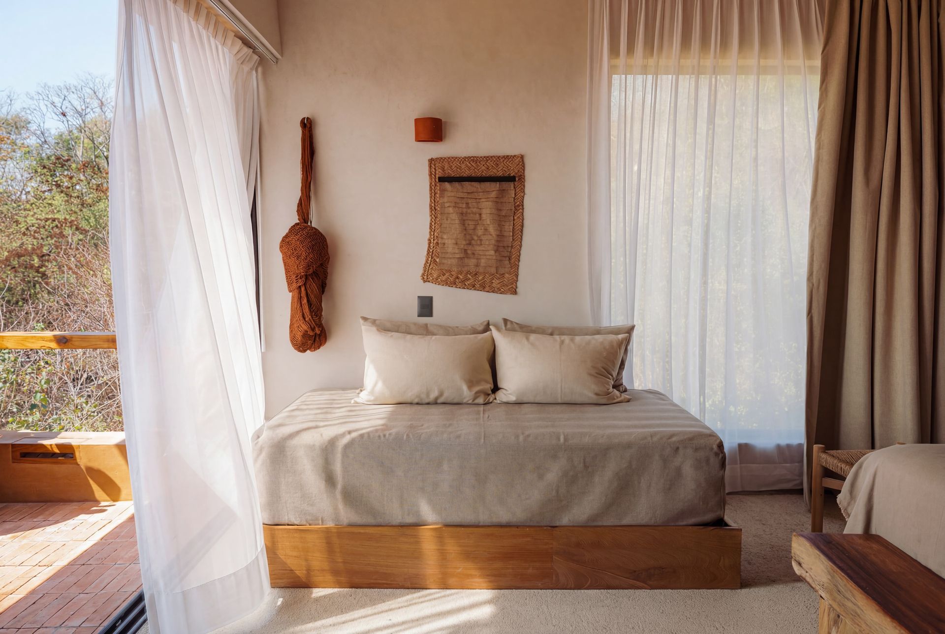 Sunlit bedroom with a beige bed, sheer curtains, woven wall decor, and a view to the outdoors.