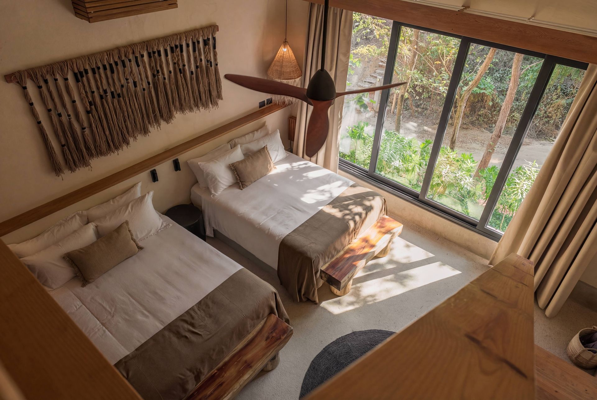 Small bedroom with two twin beds, woven wall decor, and large windows overlooking greenery.