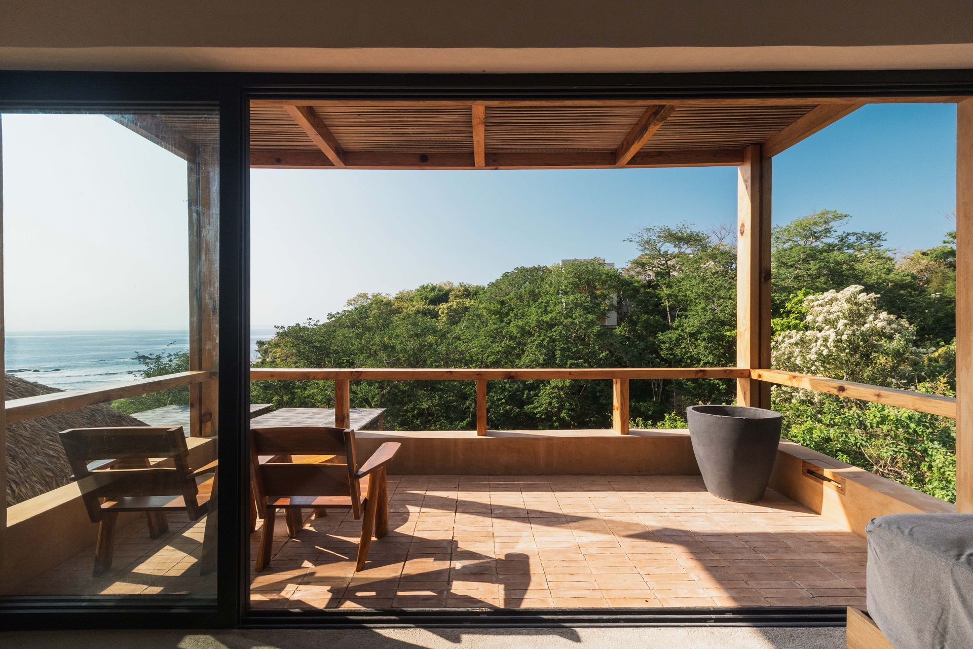 Coastal patio with wooden seating, potted plants, and ocean view through open sliding doors