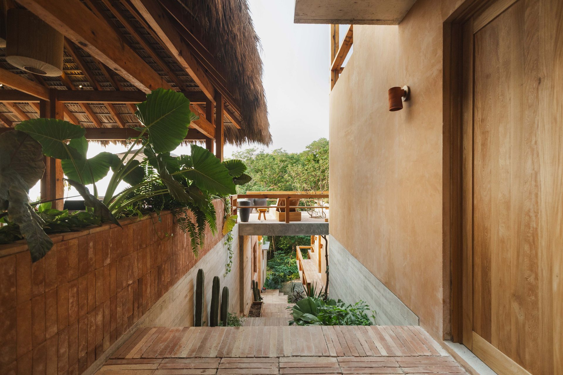 Wooden stairs between tan walls lead to a tropical courtyard with lush green plants and a covered balcony.