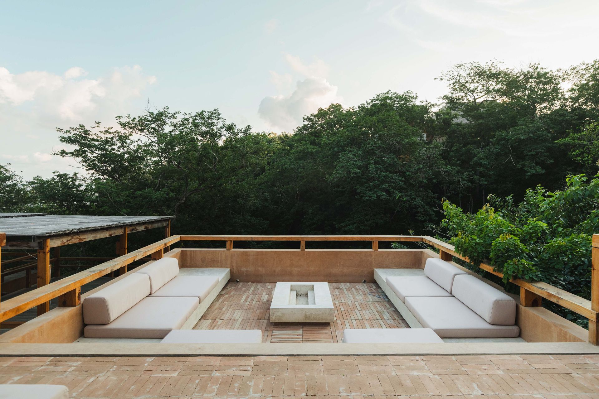 Outdoor terrace with white seating around a central table, overlooking dense green trees under a bright sky