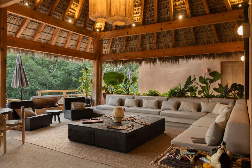 Open-air thatched lounge with low sofas, central fire pit, and wooden ceiling overlooking greenery