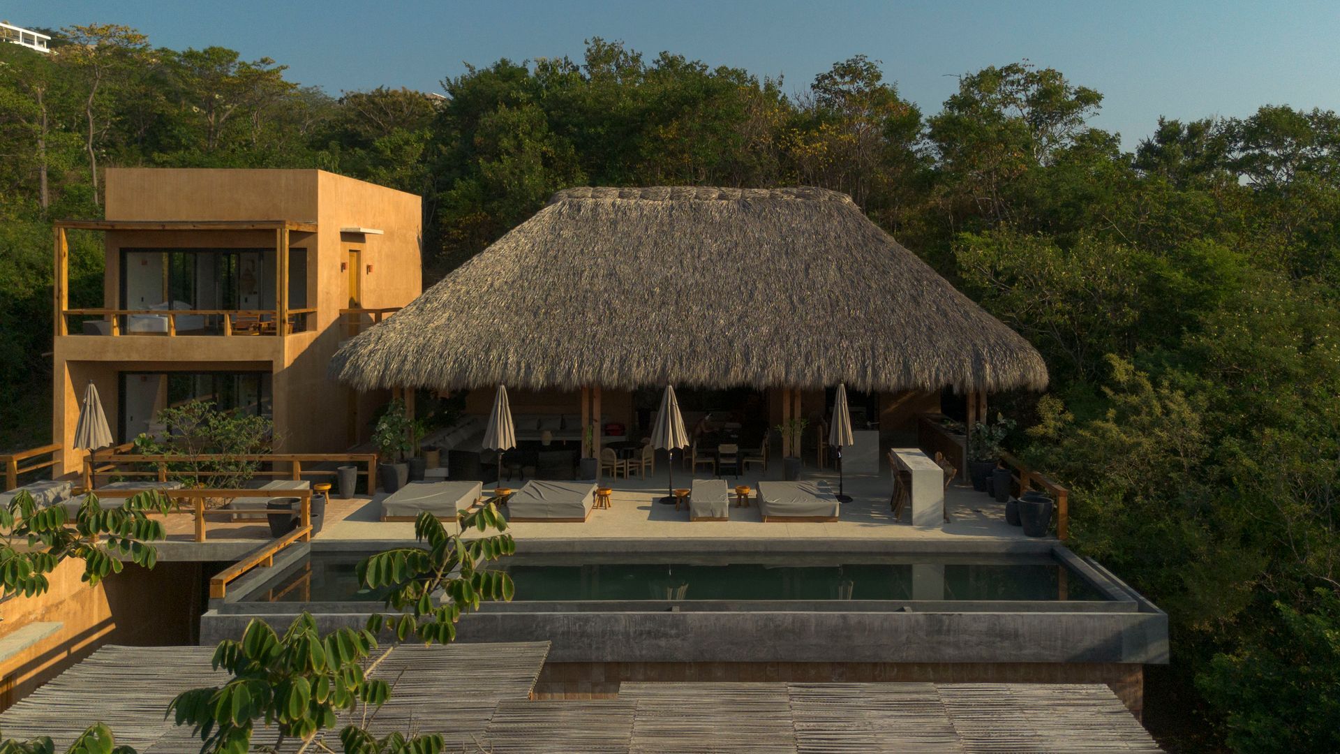 Tropical villa with thatched roof, terrace, and infinity pool amid lush greenery at sunset