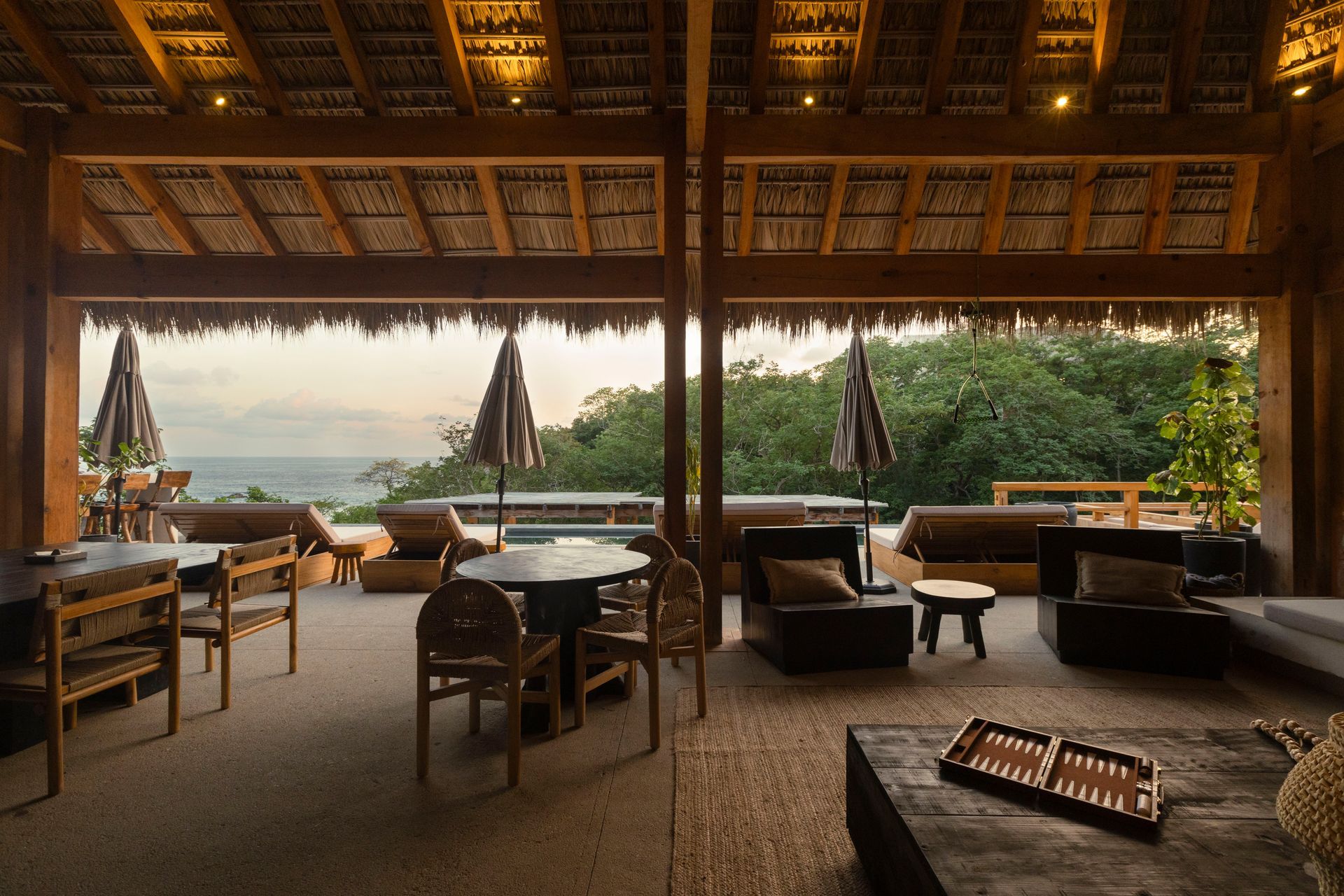 Open-air restaurant with thatched roof, tables, and lounge seating overlooking a lush tropical landscape.