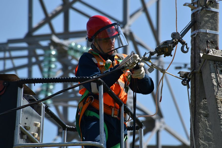 A worker in a high-visibility vest and red hard hat with a face shield repairs equipment on a utility pole from a lift.