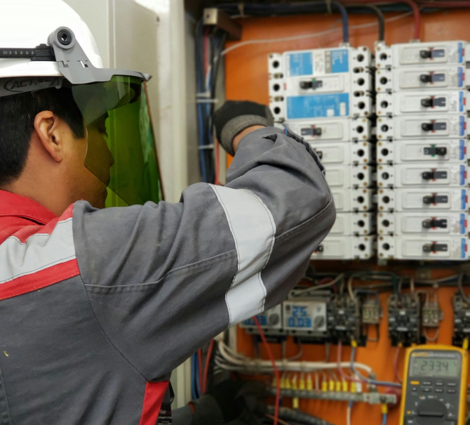 An electrician in safety gear inspects an open electrical panel with a multimeter displaying a reading.
