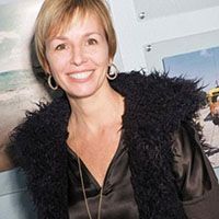 Woman with short blonde hair smiles, wearing black furry vest and jewelry.