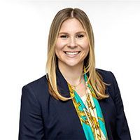Woman smiles, wearing a navy blazer, teal and gold patterned scarf, and a white shirt.