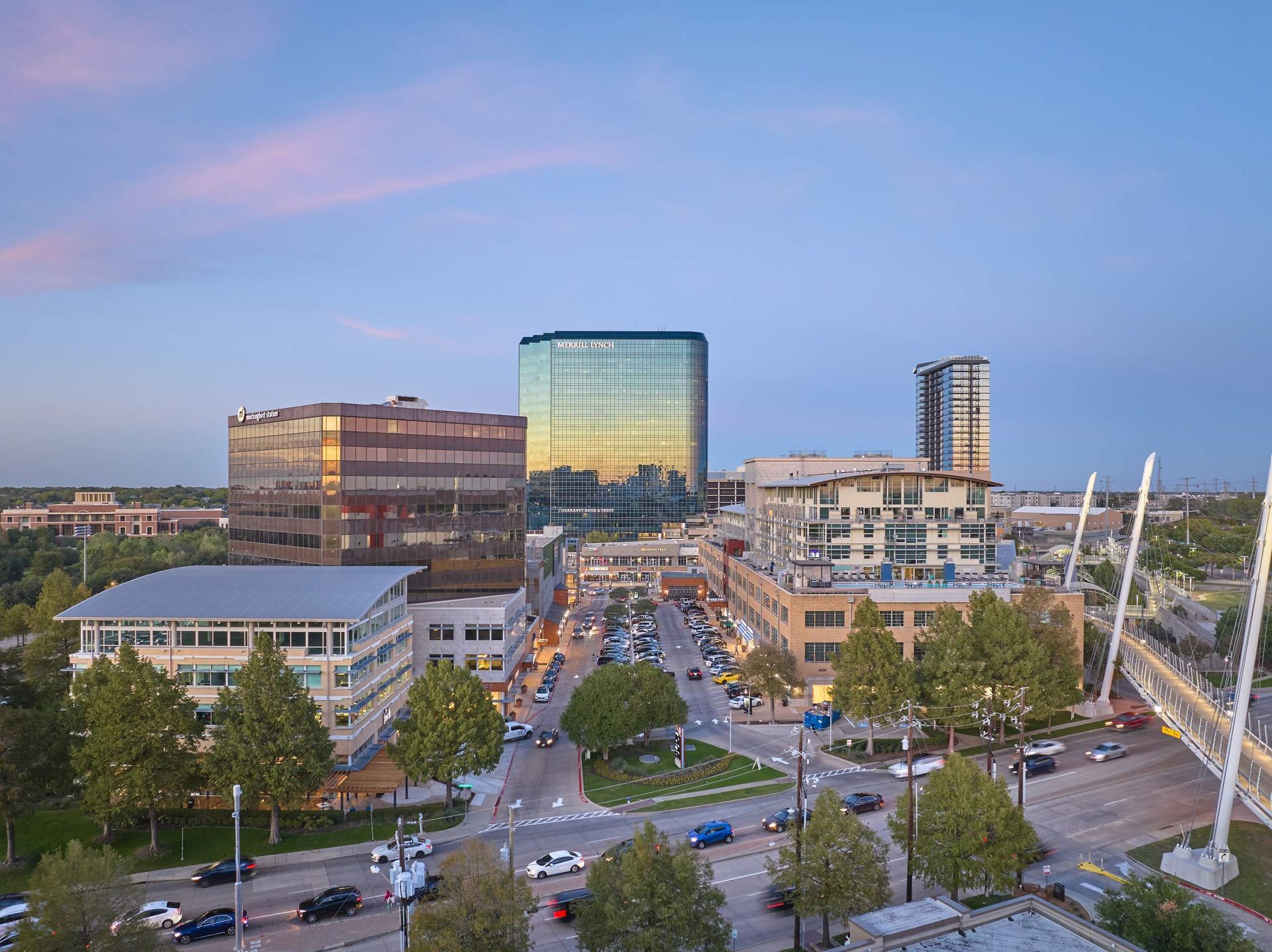 City skyline at dusk with buildings, roads, trees, and pedestrian bridge.
