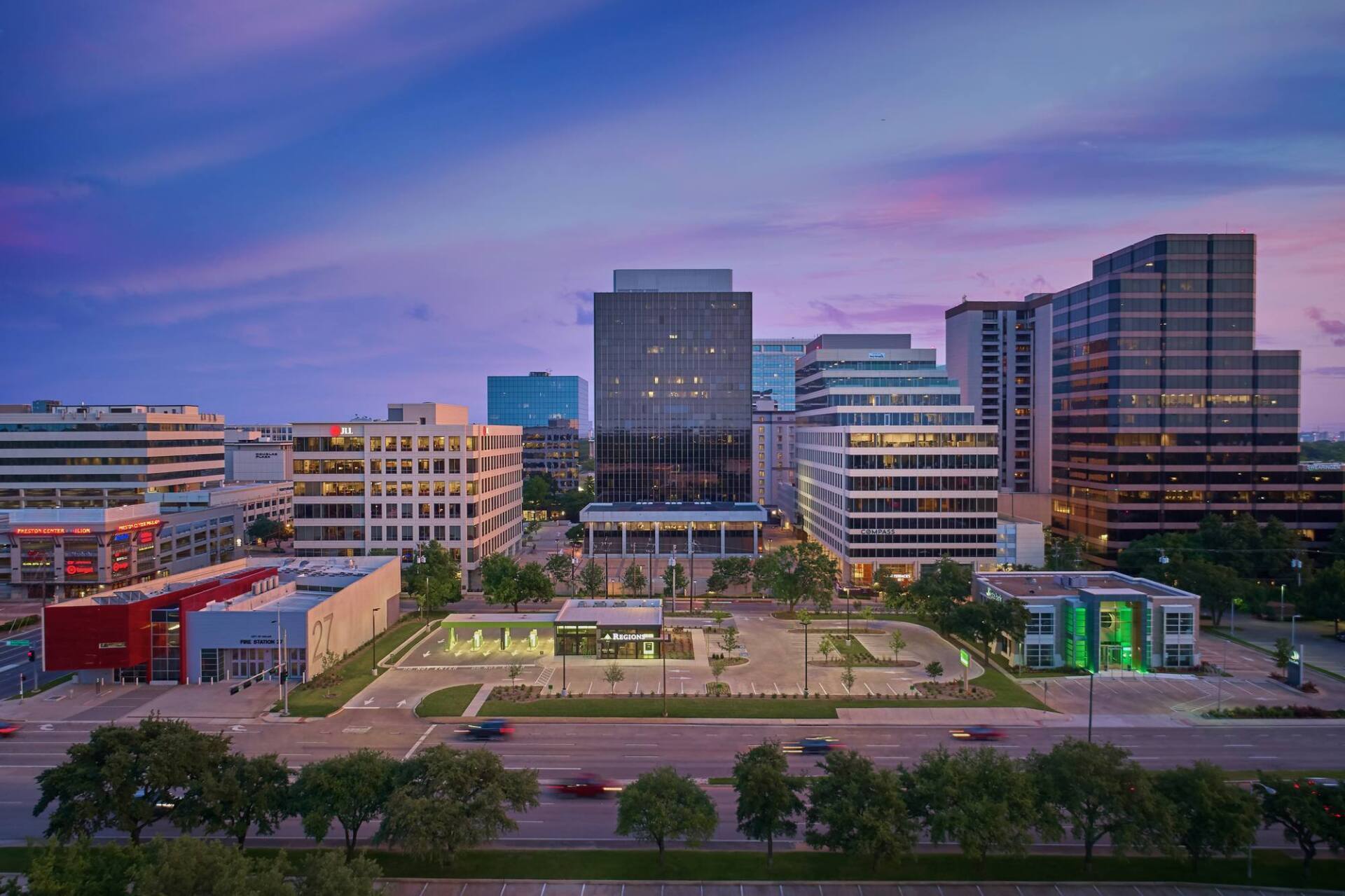 City skyline at dusk with blue and pink sky, modern office buildings, green space, and road with cars.