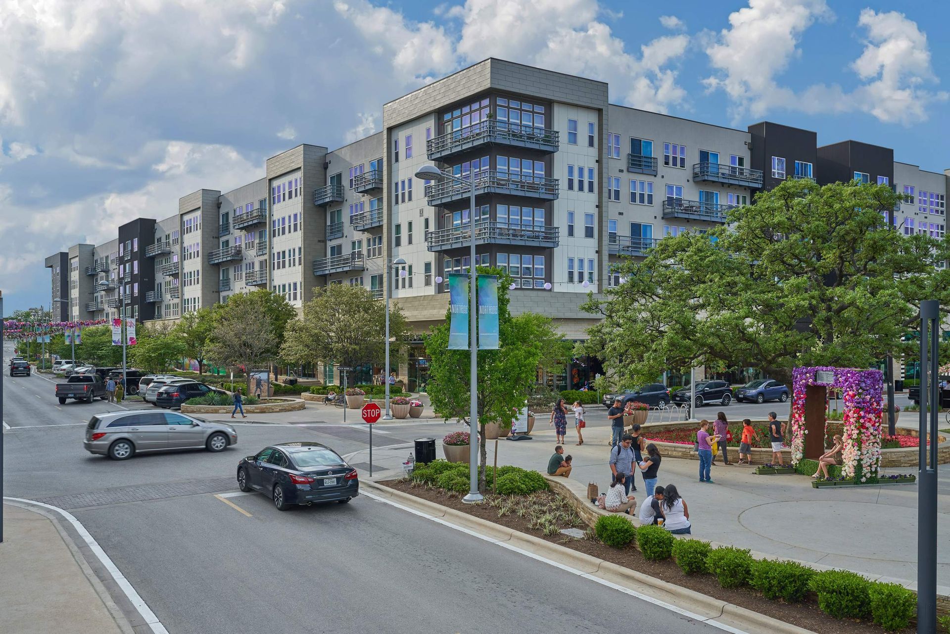 Multi-story apartment building with cars on street and pedestrians in outdoor plaza.