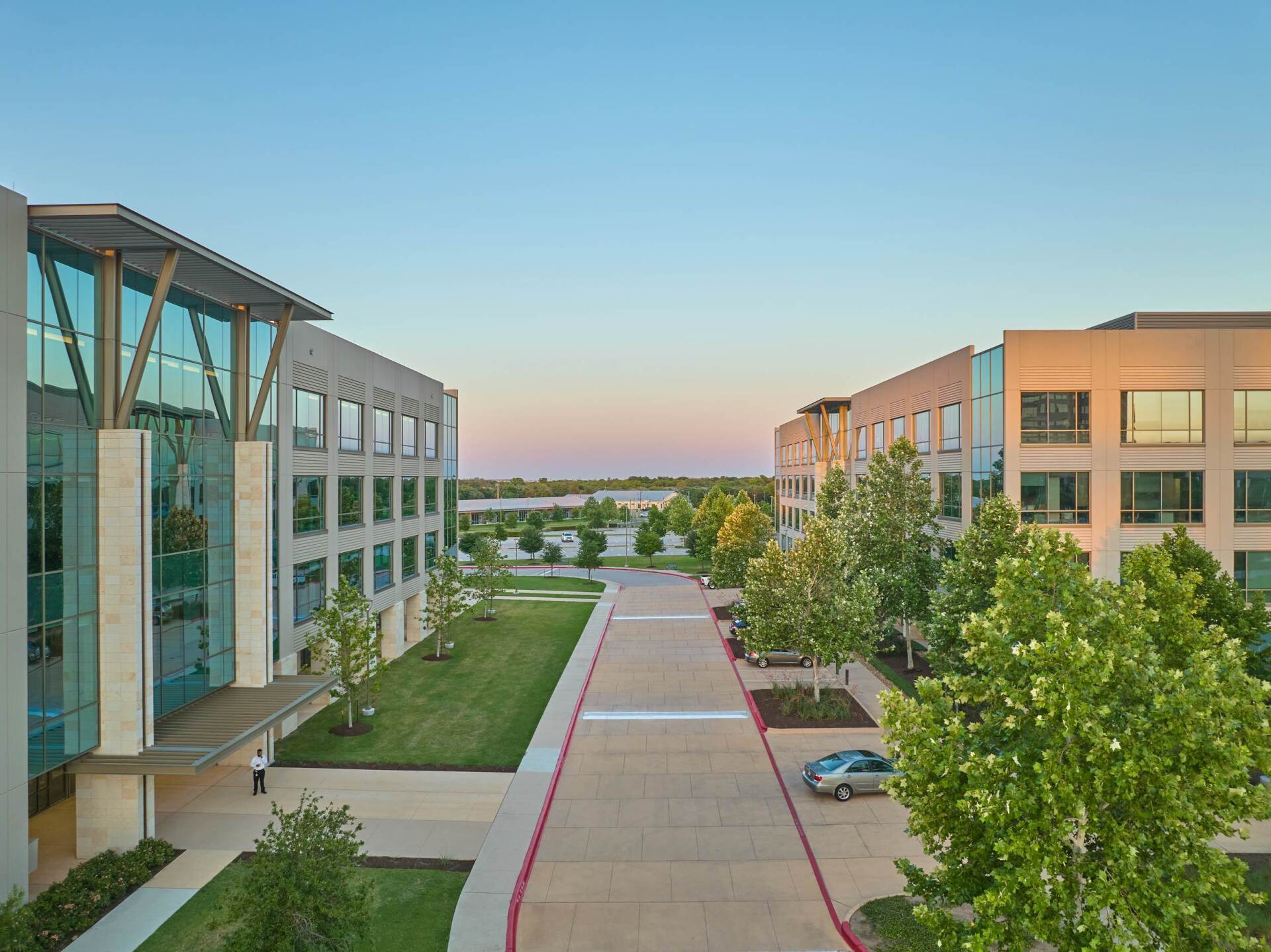 Buildings with glass and stone facade, green lawn and trees, concrete path, and a car parked on the right.