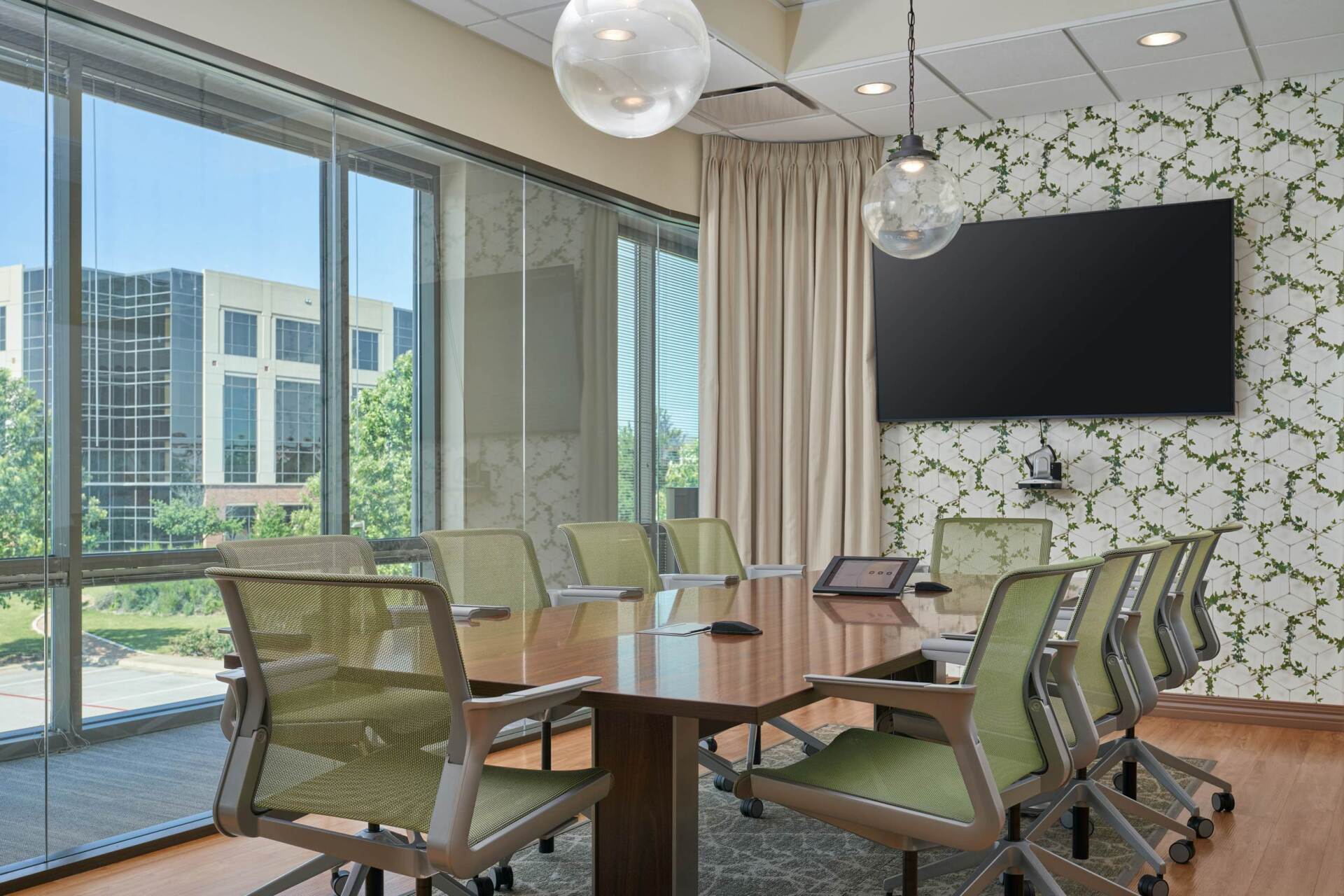Conference room with large windows overlooking a building. A long table with chairs faces a screen on patterned wallpaper.