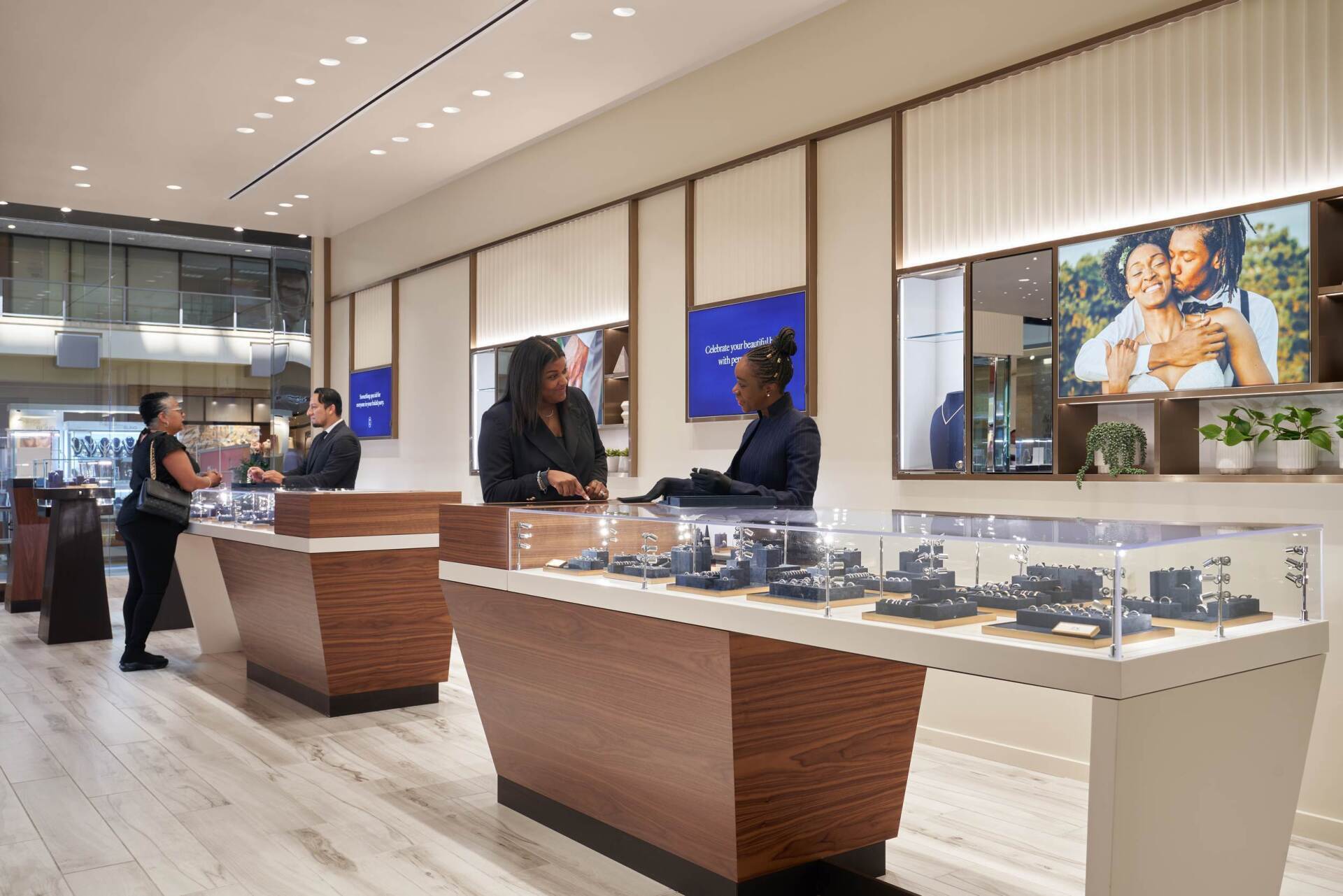 Jewelry store interior with customers and staff. Display cases, neutral colors, and bright lighting.