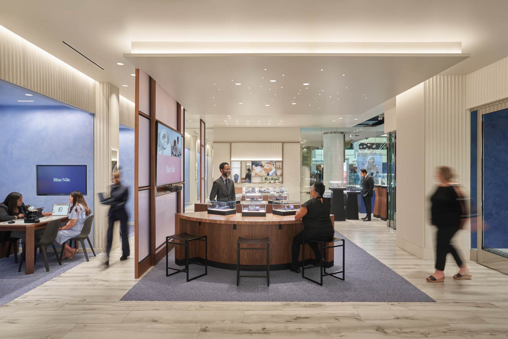 Interior of a jewelry store with customers and staff. A central counter and seating area.