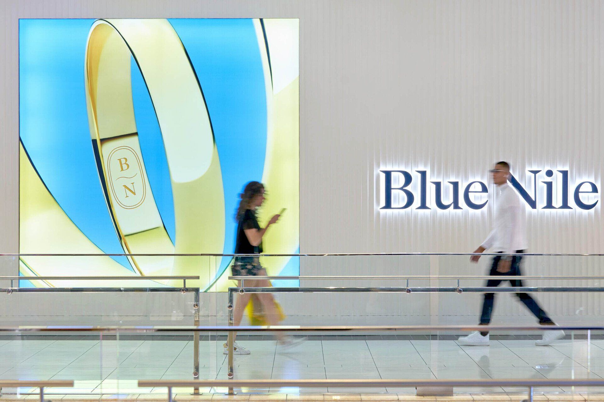 People walk past a Blue Nile store with a glowing logo and ribbon art on the wall.