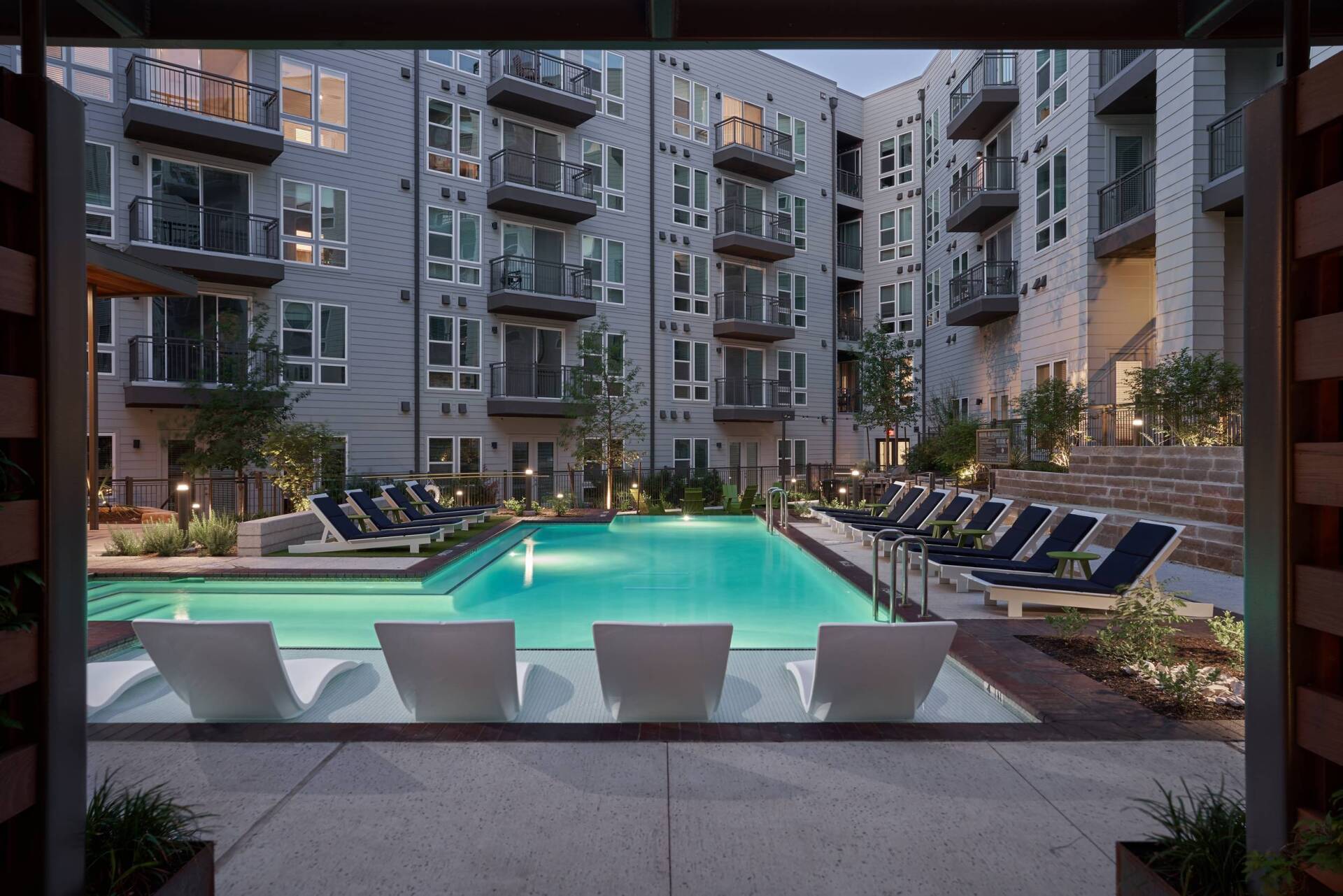 Pool area in a modern apartment complex; pool surrounded by lounge chairs, building in background.