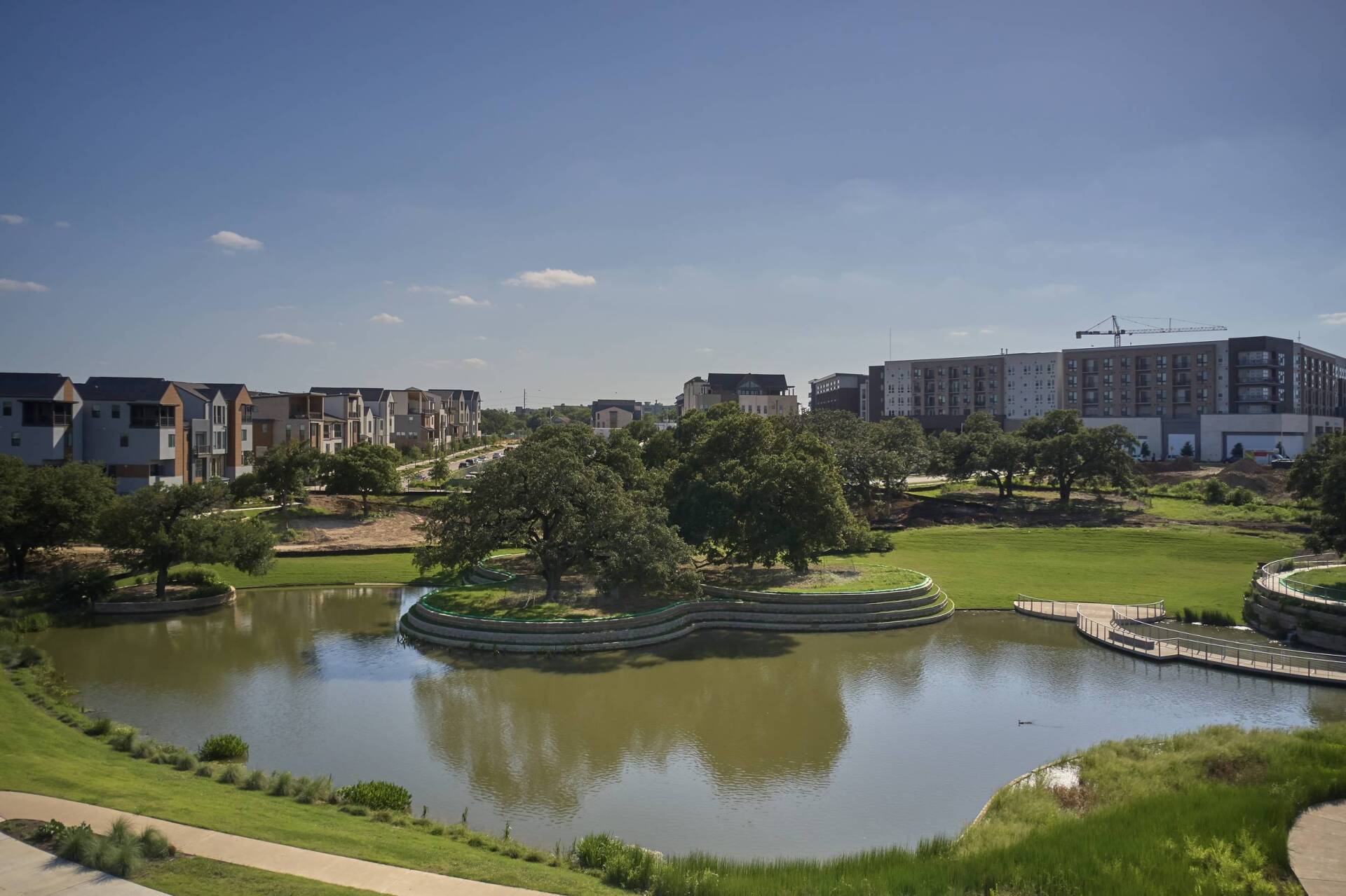 Lakeside park with houses and apartments in background under a bright blue sky. Green grass, trees, and water.