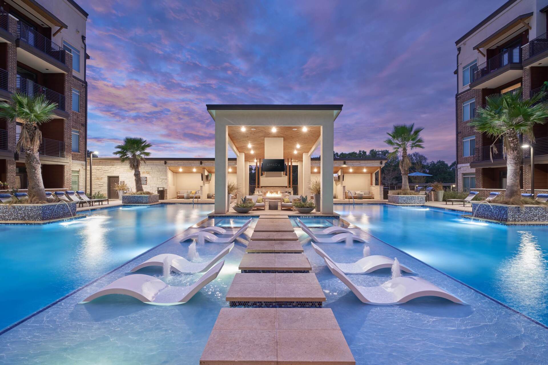 Swimming pool with lounge chairs, stone path, and covered seating area, flanked by apartment buildings at dusk.