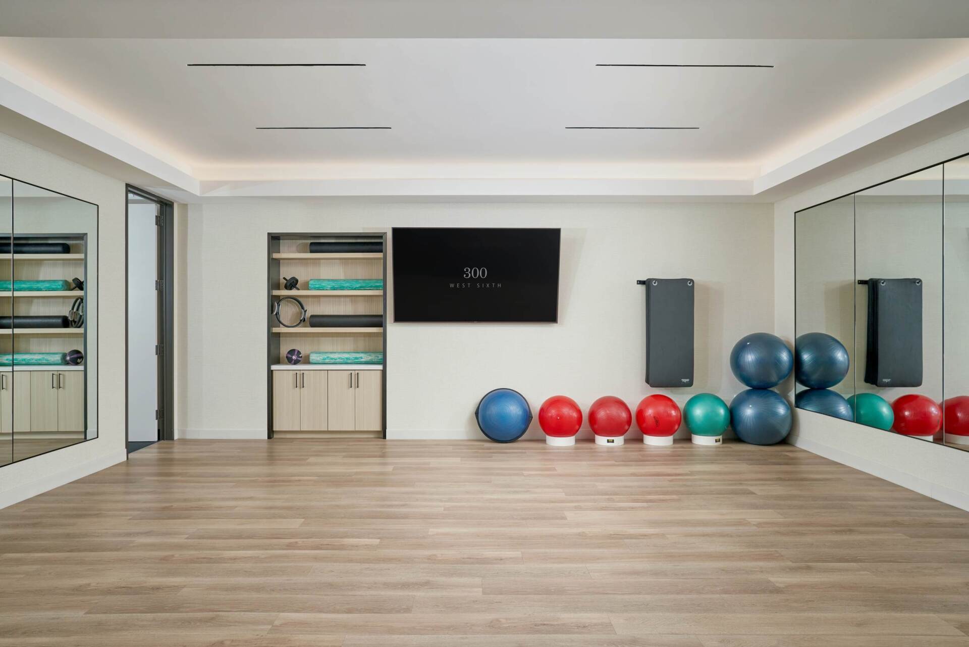 Empty exercise room with wooden floor, mirrors, exercise balls, and a TV.