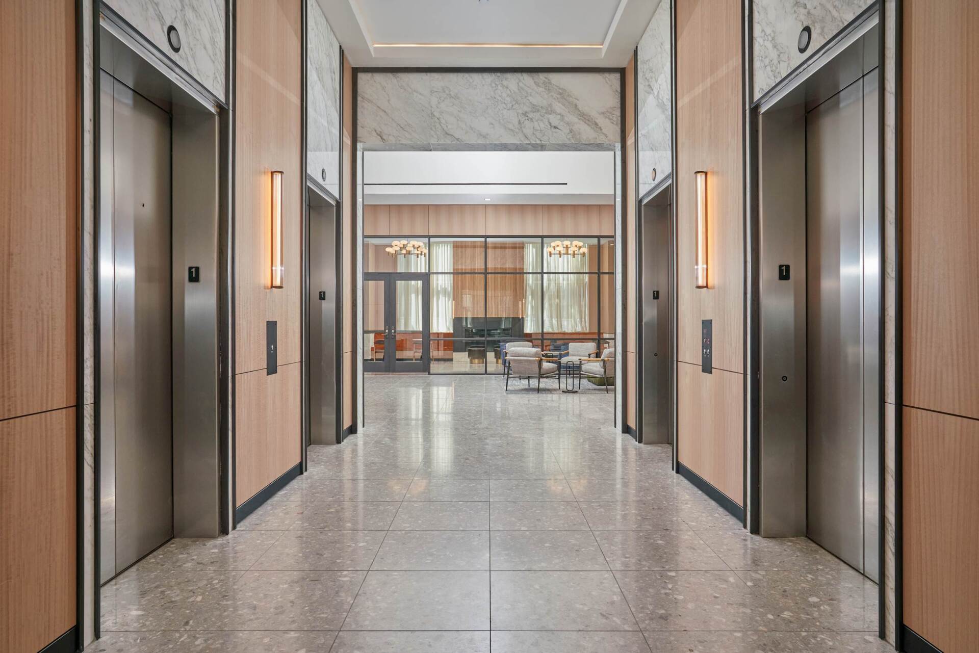Hallway with two elevators. Light wood paneling, terrazzo floor, and a seating area visible at the end.
