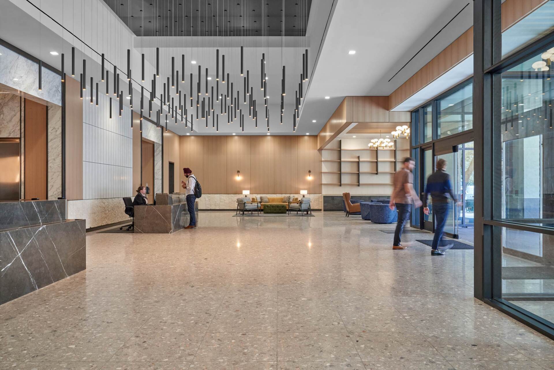Spacious lobby with terrazzo floor. People enter and check in. Modern lighting fixtures and reception area.